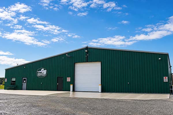 Image of a large green industrial warehouse with a white roll-up door, small side doors, and a company logo, set against a bright blue sky with scattered clouds.