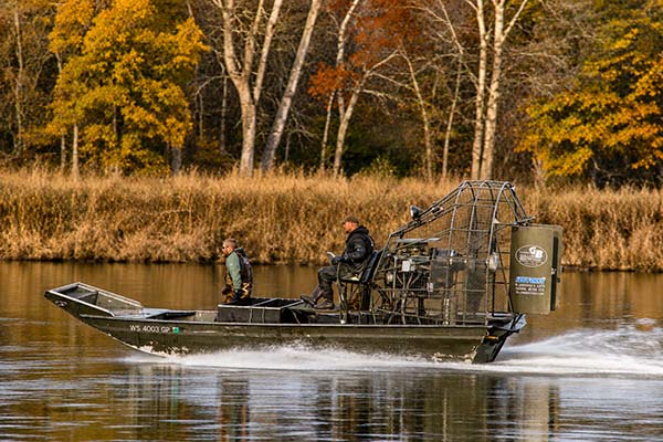 A sleek, black high-speed boat with a protective canopy navigating a calm river, surrounded by trees with vibrant fall foliage, showcasing outdoor adventure and watercraft activity.