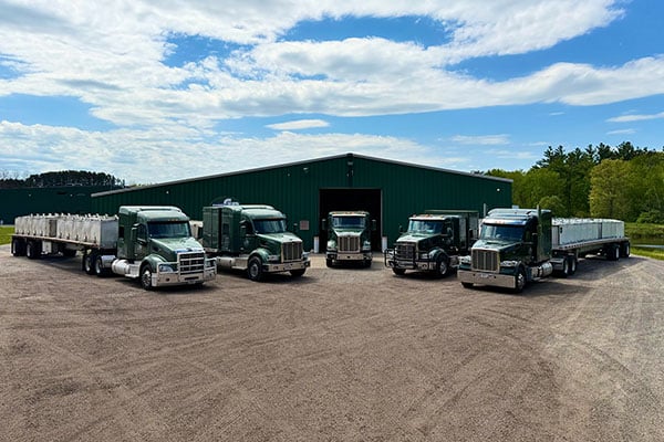 Group of five large commercial trucks parked in front of a green industrial warehouse, showcasing Gollon Brothers' fleet of heavy-duty trucks for logistics and transportation services.
