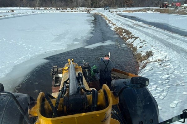 An operator using a yellow excavator to remove ice from a frozen river or canal during winter, with snow-covered banks and a cloudy sky in the background.